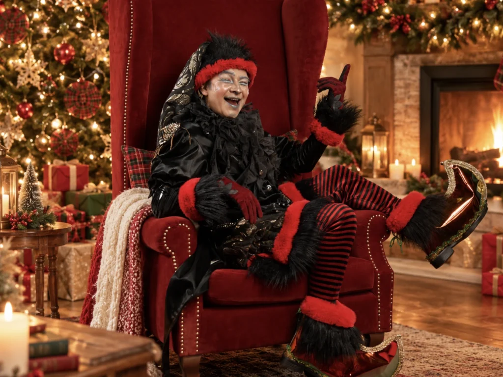 Peter Grunchy, duende vestido de negro y rojo, sentado en un sillón rojo junto a un árbol de Navidad y una chimenea